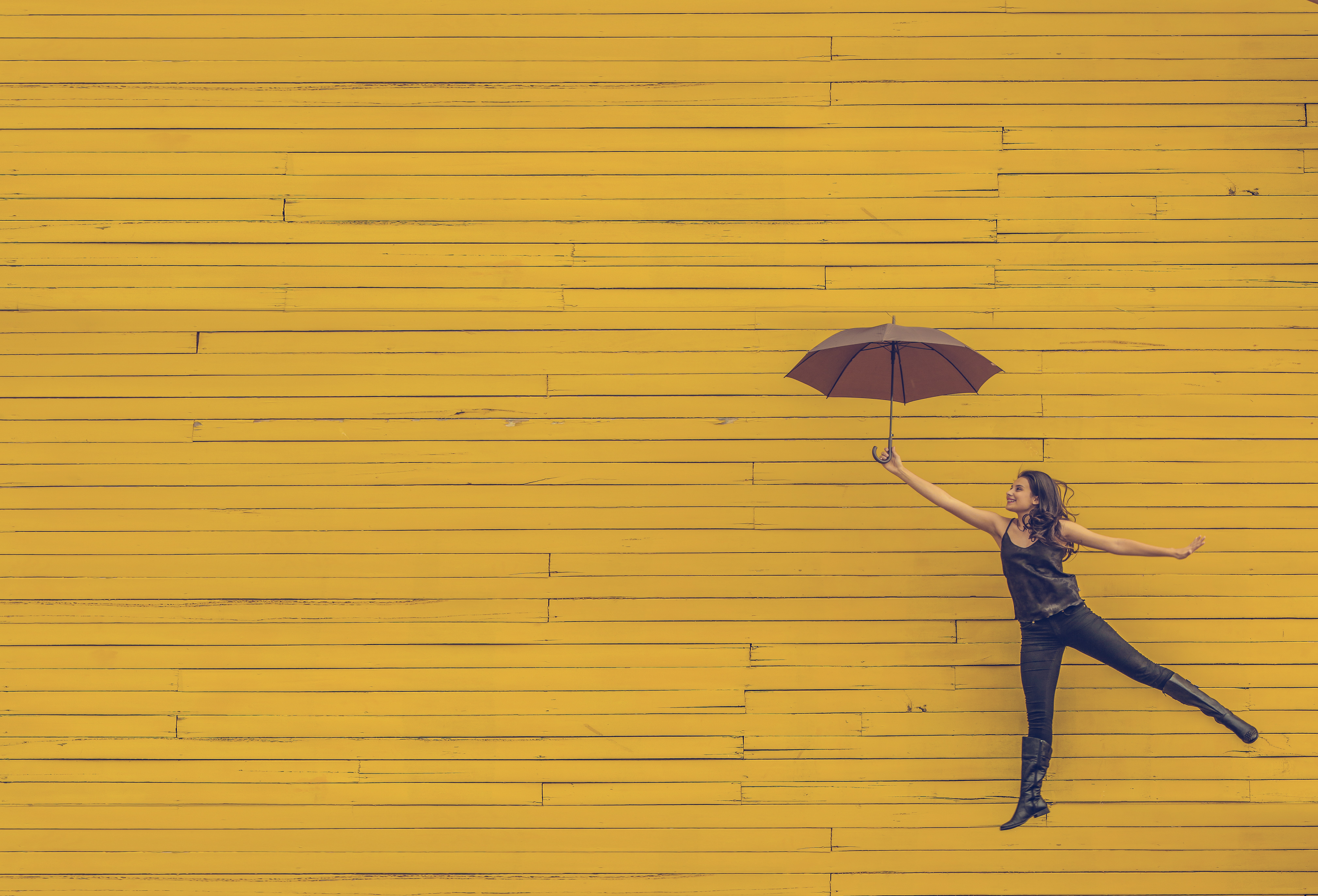 Frau springt vor gelber Wand mit einem Regenschirm in der Hand in die Luft